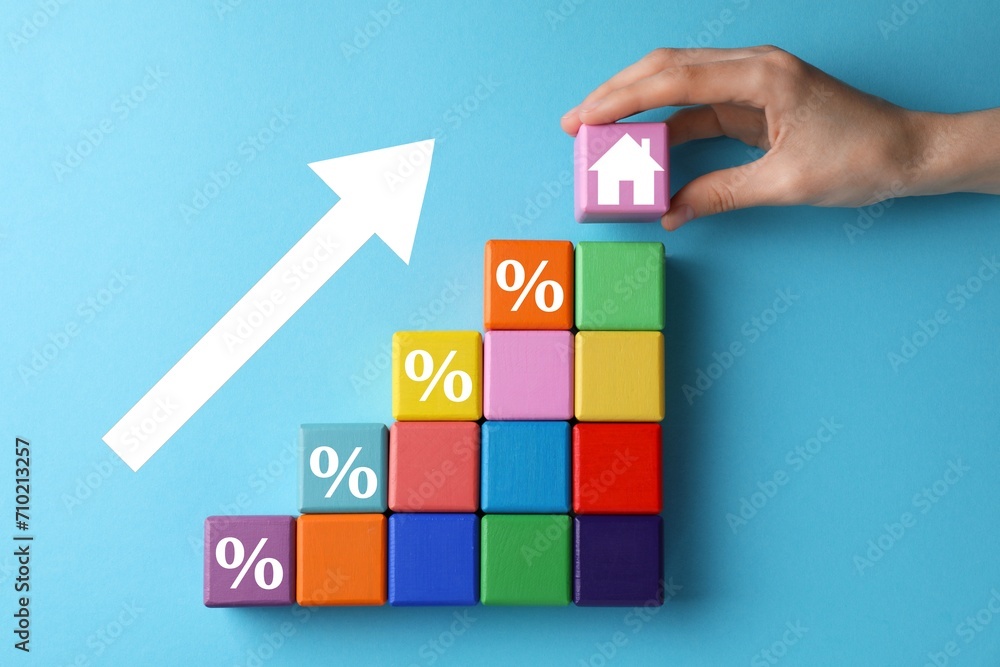 Mortgage rate rising illustrated by upward arrow. Woman putting cube with house icon near other ones on light blue background, top view Mortgage rate rising illustrated by upward arrow. Woman putting cube with house icon near other ones on light blue background, top view