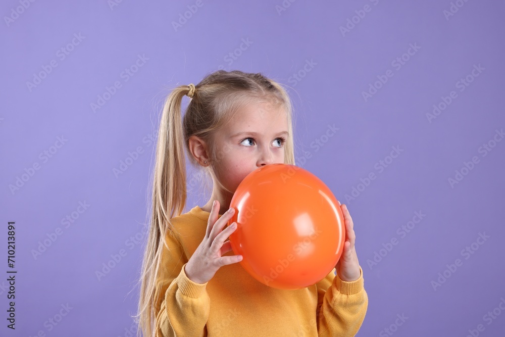 Cute little girl inflating orange balloon on violet background Cute little girl inflating orange balloon on violet background