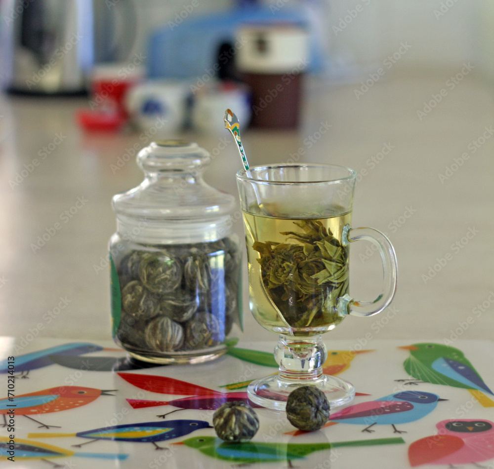 Glass of flowering tea and a jar with flowering tea balls on the place mat with birds pattern Glass of flowering tea and a jar with flowering tea balls on the place mat with birds pattern