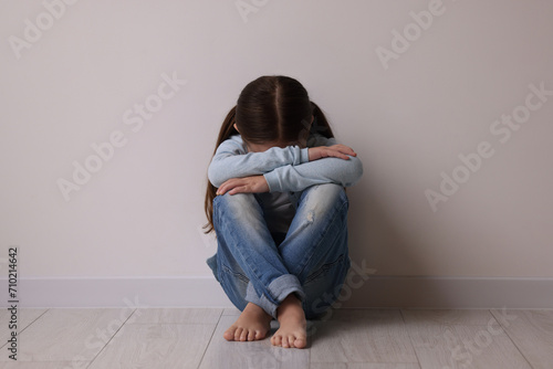 Child abuse. Upset little girl sitting on floor near light wall indoors photo
