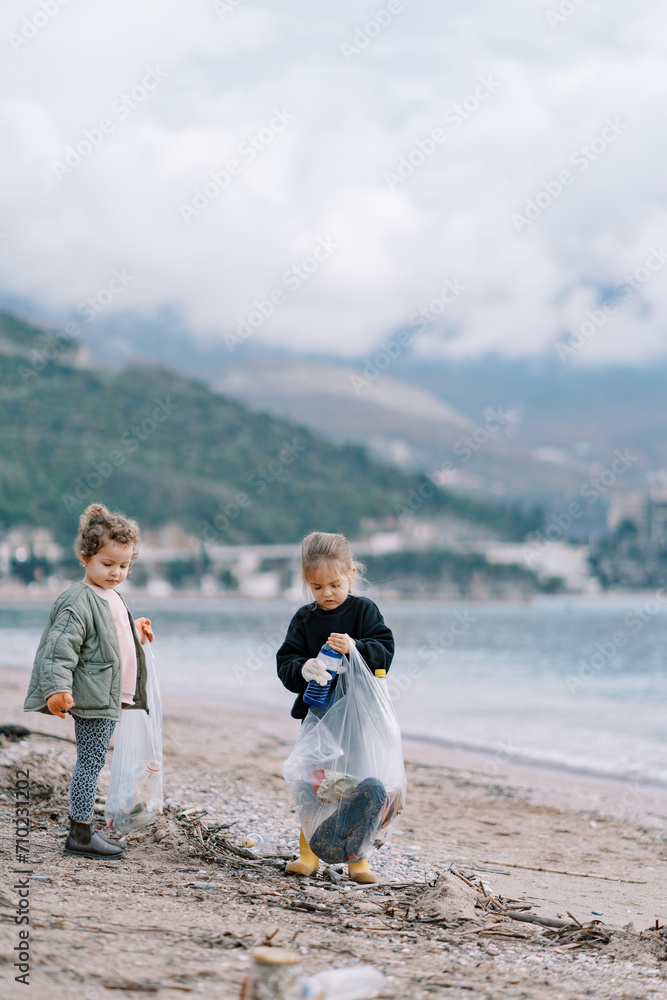 Little girls collect plastic bottles in garbage bags while walking along the sandy beach