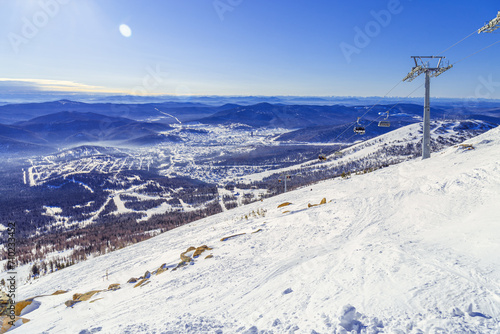 Winter nature panorama with Sheregesh ski resort in Altai, Russia, picturesque nature, sun flare on blue sky, white snow slopes and ski track, top view with range mountains, forest, perspective