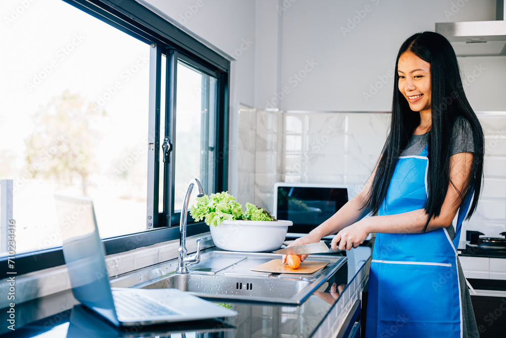 Obraz premium A woman enjoys cooking in her kitchen seeking recipes on her laptop. Portraying the joy and convenience of technology in meal preparation and culinary endeavors.