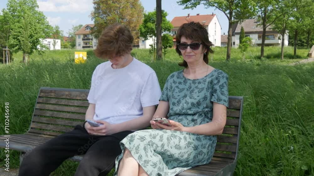 An adult mother and a teenager son sit on a bench in a summer park and look at a smartphone, press the screen, hold the phone in their hands An adult mother and a teenager son sit on a bench in a summer park and look at a smartphone, press the screen, hold the phone in their hands