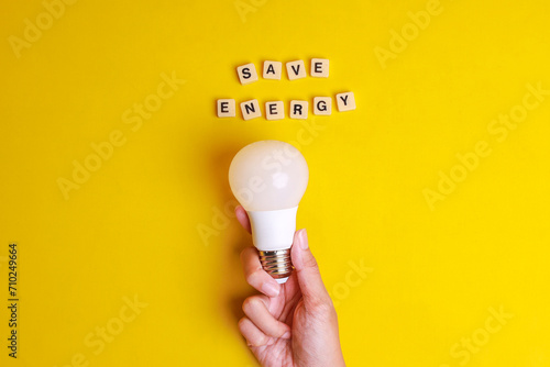 Save Energy words written on wooden blocks with hand holding white light bulb on yellow background. Concept of saving energy by using LED lightbulb photo