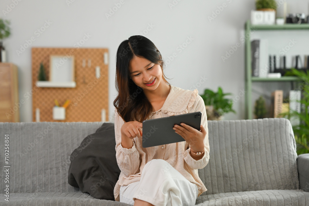 Smiling young woman in casual clothes using digital tablet on couch at cozy living room Smiling young woman in casual clothes using digital tablet on couch at cozy living room