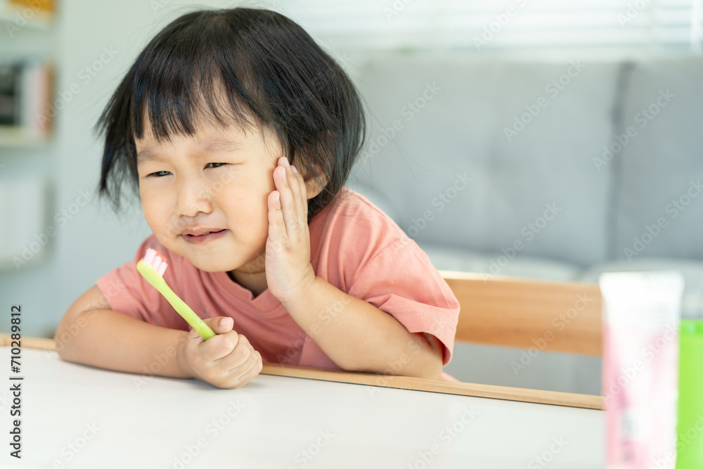 little asian girl presses hand to cheek, suffers from pain in tooth. Teeth decay, dental problems, child emotions and facial expression, oral health care, reducing sweets, fluorine coating.