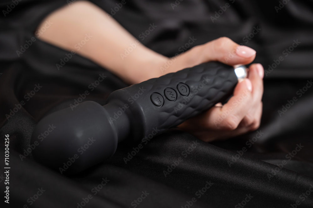 A woman is holding a black vibrator while lying on a black silk sheet.