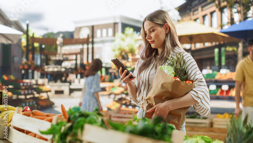 Young Beautiful Customer Shopping for Fresh Seasonal Fruits and Vegetables, Using Smartphone to Browse Internet on the Move. Female Holding a Sustainable Paper Bag with Ecological Local Farm Produce