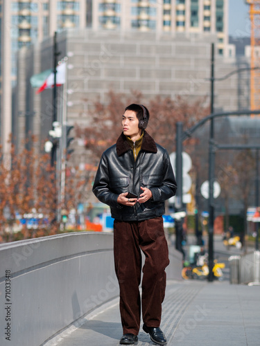Portrait of handsome Chinese young man with black leather jacket wearing headset holding mobile phone posing with modern city street background in sunny winter day, male fashion, cool Asian young man.