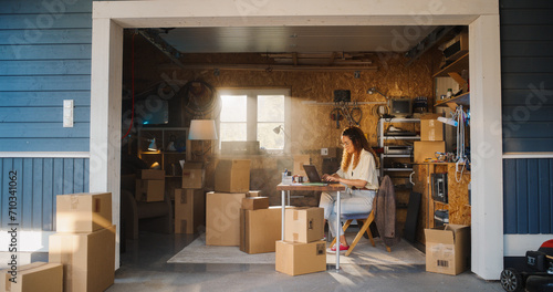 Hispanic Businesswoman Using Laptop Computer In Garage. Female Small Business Owner Filling Orders From Online Customers. Products In Cardboard Boxes Ready For Shipping. Entrepreneur Working At Home. photo