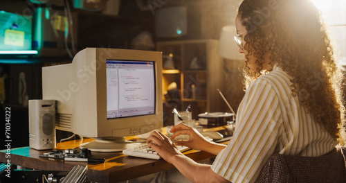 Side View Of Hispanic Female Engineering Student Writing Academic Thesis On Old Desktop Computer In Retro Garage. Future Engineer Focused On Innovative Project For University Of Technology In Nineties