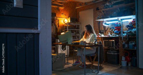 Wide Shot Of Hispanic Female Engineer Using Old Computer In Nineties Retro Garage In The Evening. Intelligent Woman Programming, Working On Innovative Portable Device, Starting Technological Company.