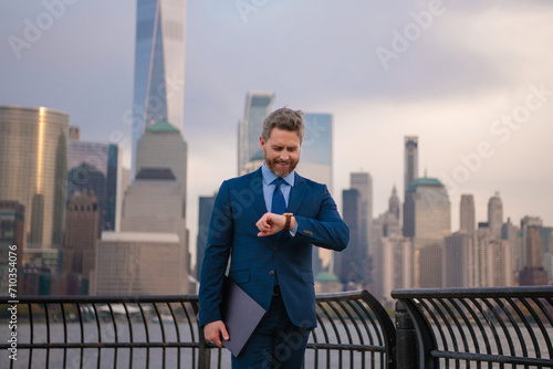 Businessman looking on watch to managing time for organization of business. Middle aged business man standing outside office using laptop. Street portrait of mature businessman.