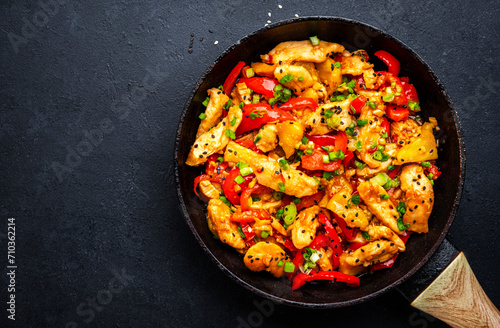Stir fry chicken slices with pineapple, paprika, green onion, soy sauce and sesame seeds in frying pan. Asian cuisine dish. Black table background, top view