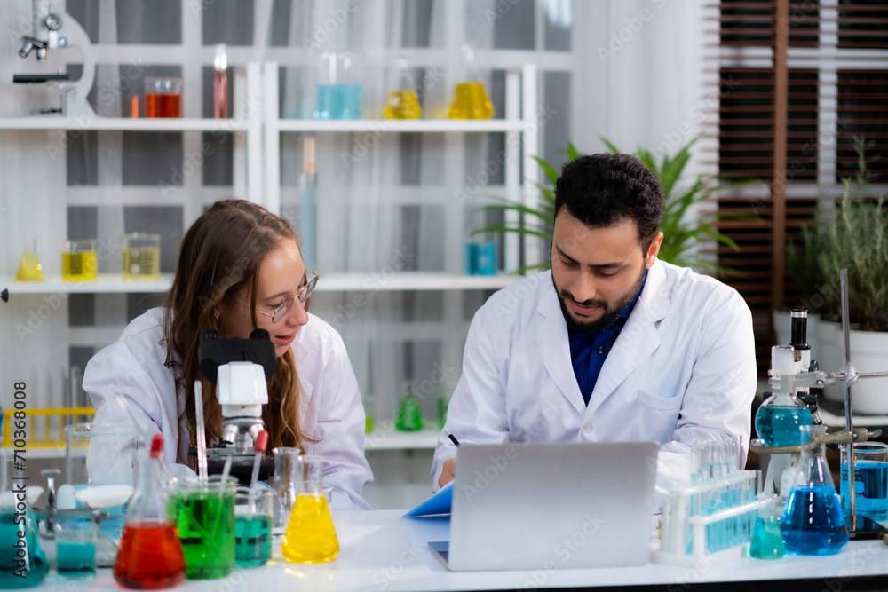 Young White Sciencetist woman and man sitting at desk with laptop and colorful liquid in experiment equipment talking about experiment result with smile of successs. Young White Sciencetist woman and man sitting at desk with laptop and colorful liquid in experiment equipment talking about experiment result with smile of successs.