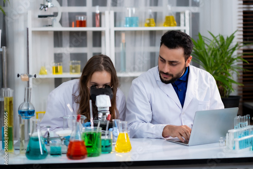 Young White Sciencetist woman looking into microscope while her co-worker looking at her waitting for result of experiment in Lab room.