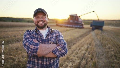 Happy bearded farmer in cap looking at camera. Farmer on agricultural wheat field with combine harvester and dump truck on background. Harvesting, farming at sunset. Food production, agribusiness. photo