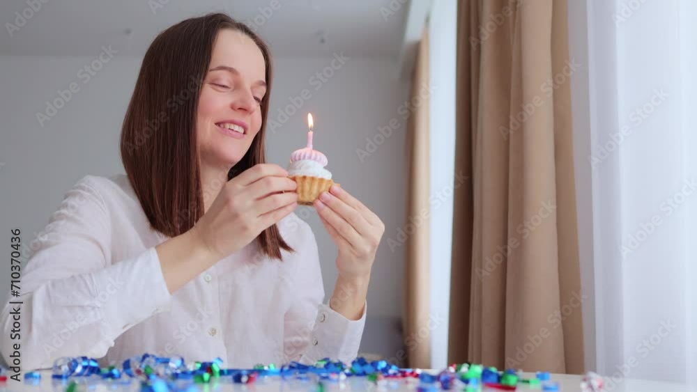 Making wish on birthday. Caucasian young adult woman sitting at table with festive cake blowing burning candle in light room interior enjoying her party alone Making wish on birthday. Caucasian young adult woman sitting at table with festive cake blowing burning candle in light room interior enjoying her party alone