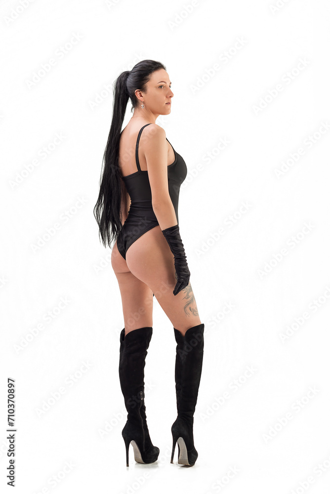 A gorgeous slim, tanned brunette with a perfect body in a black bodysuit and boots poses against a white background in the studio. Hair removal. A gorgeous slim, tanned brunette with a perfect body in a black bodysuit and boots poses against a white background in the studio. Hair removal.