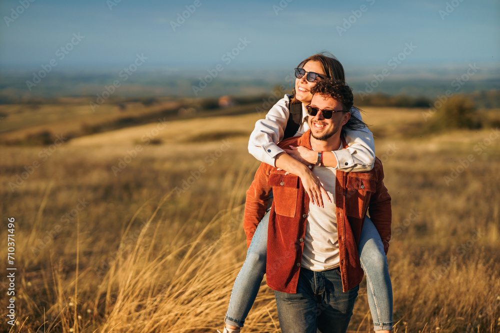 A boy is holding a girl on his back while walking through golden yellow meadow on a sunny day. They are wearing a sunglasses and smiling.