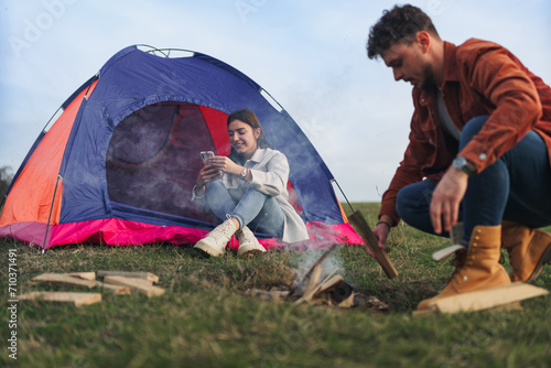 A girl is looking something on her phone in front of a tent, while a boy is making a fire on a golden yellow meadow.
