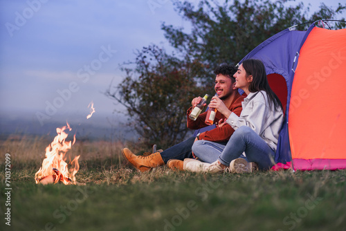 A happy couple is sitting and drinking a beverage in front of a blue tent by the campfire. They enjoy camping in nature.