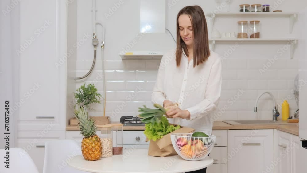 Brown haired woman wearing white shirt unpacking fruits and vegetables after buying in supermarket preparing fresh products for cooking healthy dish putting food on table in kitchen at home Brown haired woman wearing white shirt unpacking fruits and vegetables after buying in supermarket preparing fresh products for cooking healthy dish putting food on table in kitchen at home