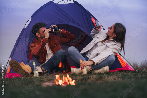 A couple is enjoying camping in nature, a boy is holding binoculars while a girl is taking a photo of him.