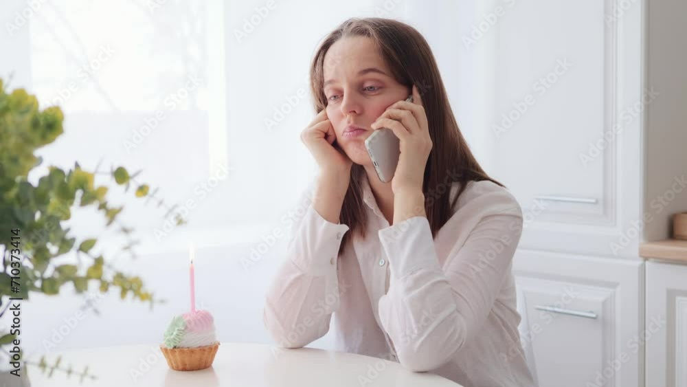 Sad depressed adult woman wearing white shirt celebrating birthday alone calling to her friend inviting to her party felling loneliness and sadness sitting at table in kitchen with cake and candle Sad depressed adult woman wearing white shirt celebrating birthday alone calling to her friend inviting to her party felling loneliness and sadness sitting at table in kitchen with cake and candle
