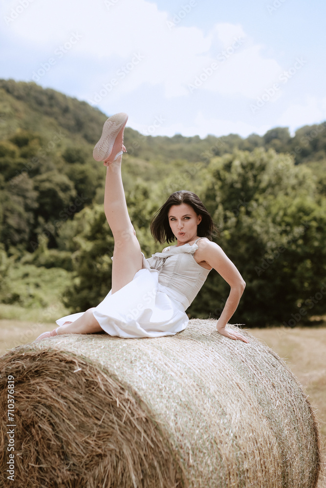 Portrait of a beautiful woman in a summer dress on a field among haystacks in summer. A young, slender girl in a white sundress poses among stacks of yellow dry grass on a sunny summer day. High quali Portrait of a beautiful woman in a summer dress on a field among haystacks in summer. A young, slender girl in a white sundress poses among stacks of yellow dry grass on a sunny summer day. High quali