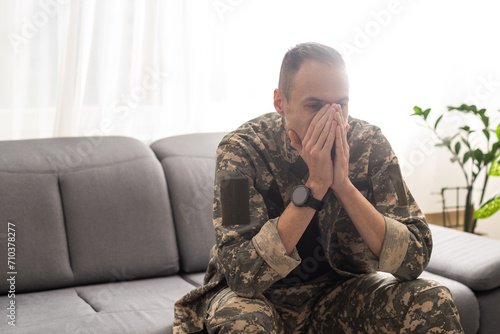 Thoughtful military man staring aside, holding palms by mouth, sitting on couch at home. Young soldier visiting psychologist, suffering from posttraumatic stress, closeup photo, copy space photo