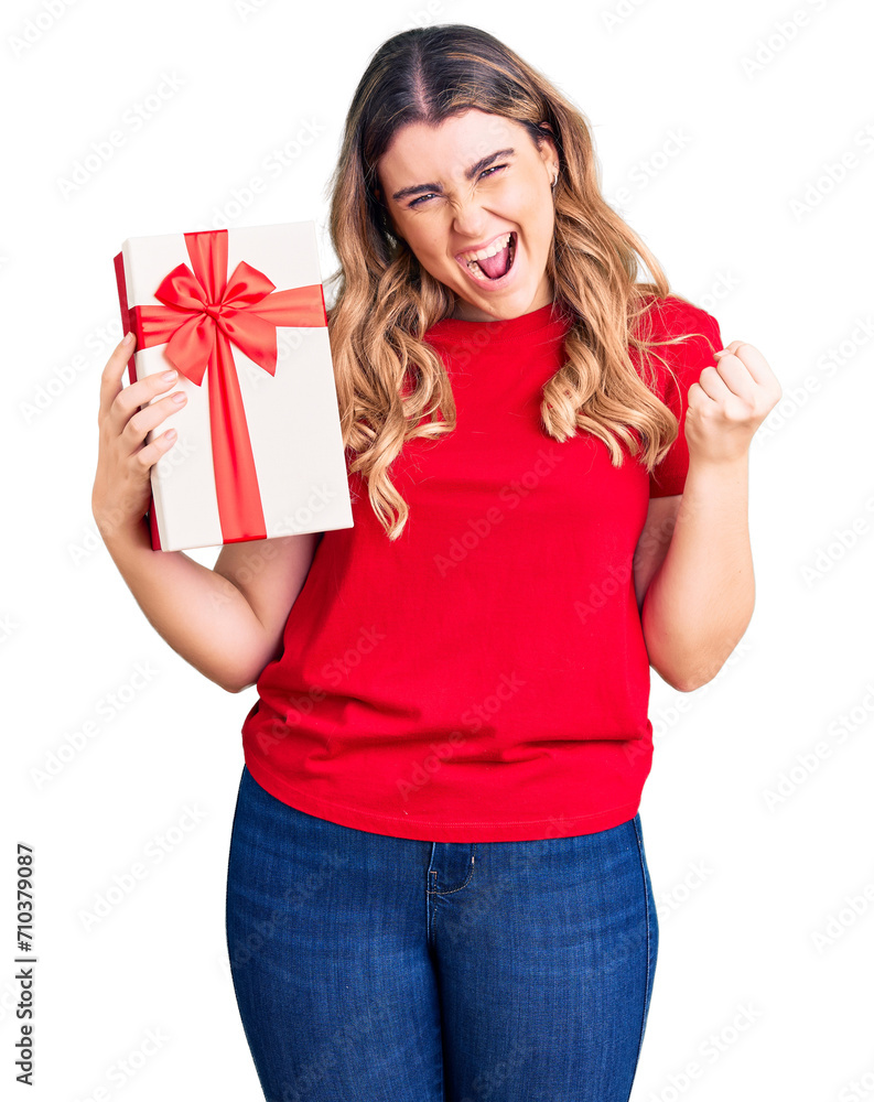 Fototapeta premium Young caucasian woman holding gift screaming proud, celebrating victory and success very excited with raised arms