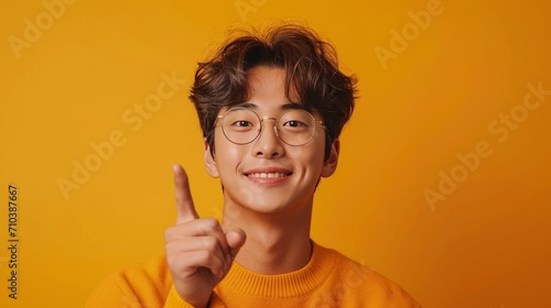young korean man Pointing playfully up against a bright yellow background. Embodying the spirit of optimism and positive energy.