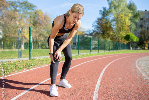 Female athlete in sportswear pausing her exercise due to a painful knee injury on a sunny outdoor track.