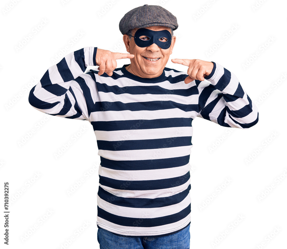 Senior handsome man wearing burglar mask and t-shirt smiling cheerful showing and pointing with fingers teeth and mouth. dental health concept. Senior handsome man wearing burglar mask and t-shirt smiling cheerful showing and pointing with fingers teeth and mouth. dental health concept.