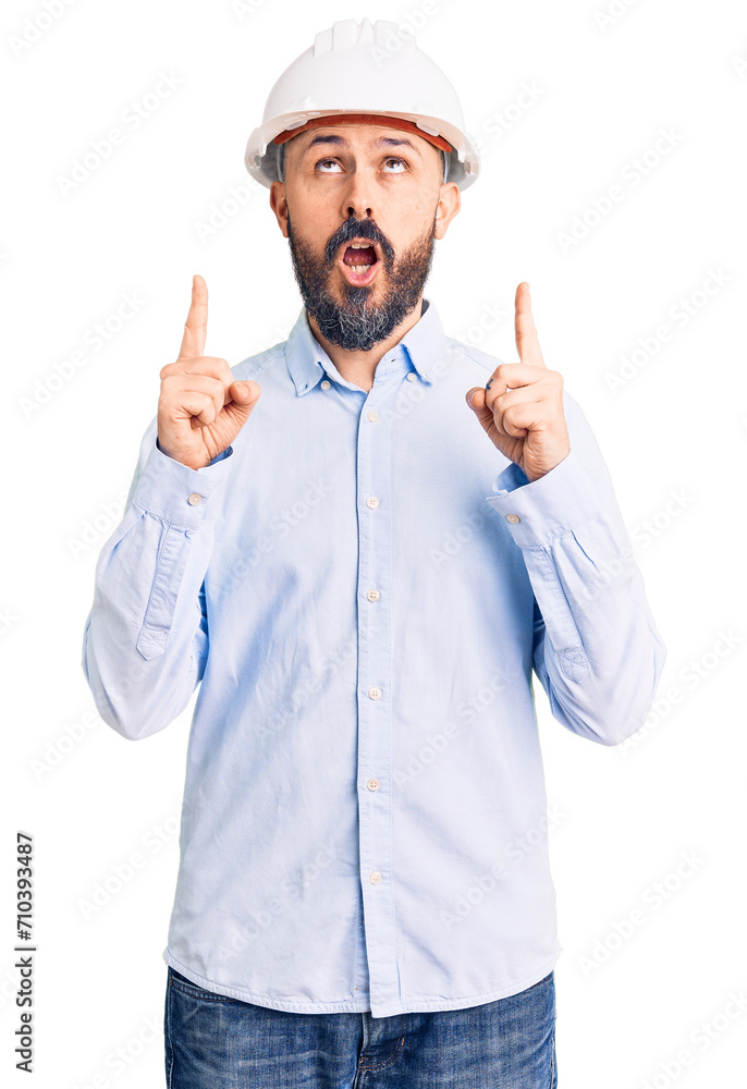 Young handsome man wearing architect hardhat amazed and surprised looking up and pointing with fingers and raised arms. Young handsome man wearing architect hardhat amazed and surprised looking up and pointing with fingers and raised arms.