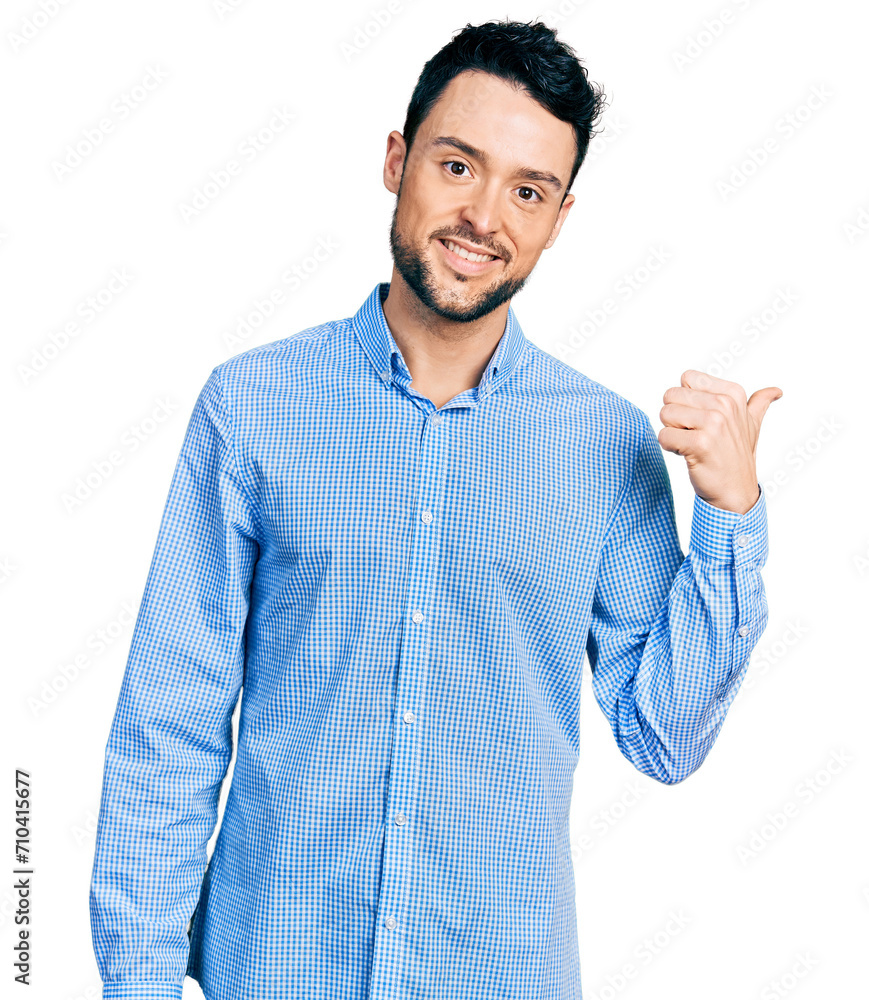 Hispanic man with beard wearing casual business shirt smiling with happy face looking and pointing to the side with thumb up. Hispanic man with beard wearing casual business shirt smiling with happy face looking and pointing to the side with thumb up.