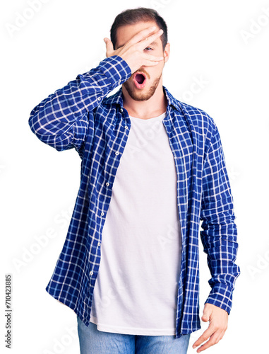 Young handsome caucasian man wearing casual clothes peeking in shock covering face and eyes with hand, looking through fingers with embarrassed expression.