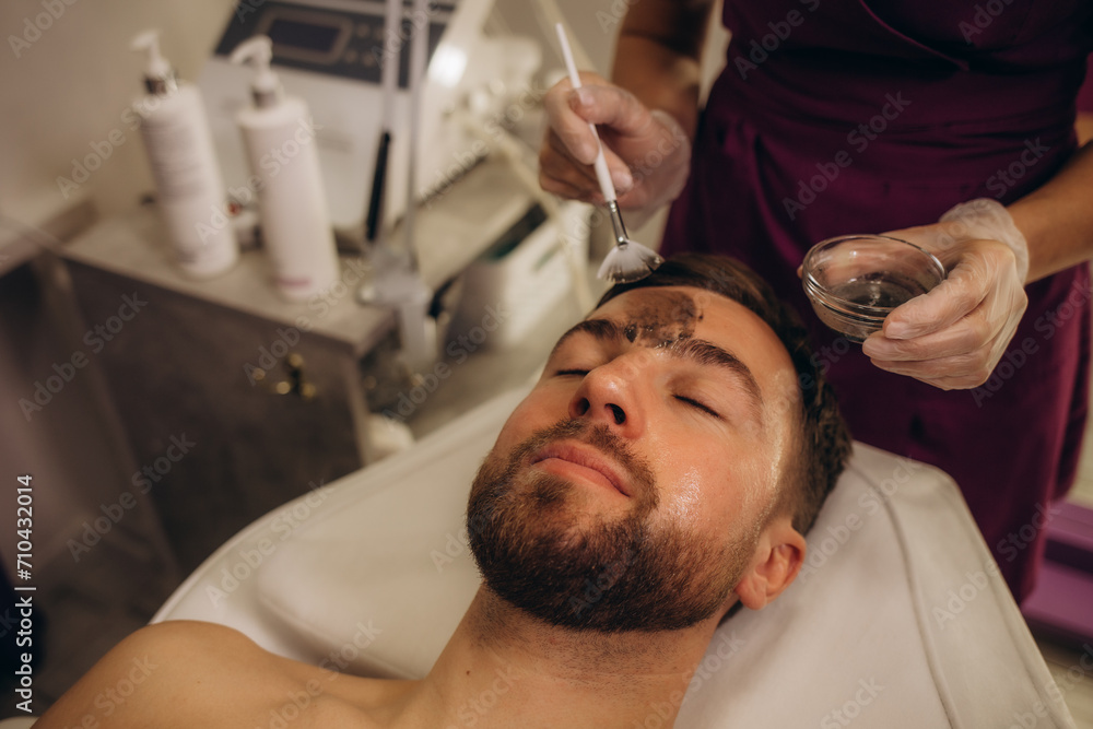 Fototapeta premium Above angle of relaxed and serene young man keeping head on rolled soft towel while beautician applying clay mask on his face in spa salon