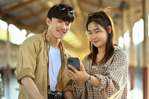 Cheerful Asian couple tourist checking timetable train schedule for trip on mobile phone