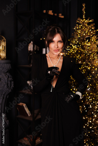 Beautiful woman in long black evening dress, celebrate New Year, Christmas Tree behind. Big lustre. Posch middle-aged lady with gorgeous jewelry of pearls. Portrait of girl in black room. 