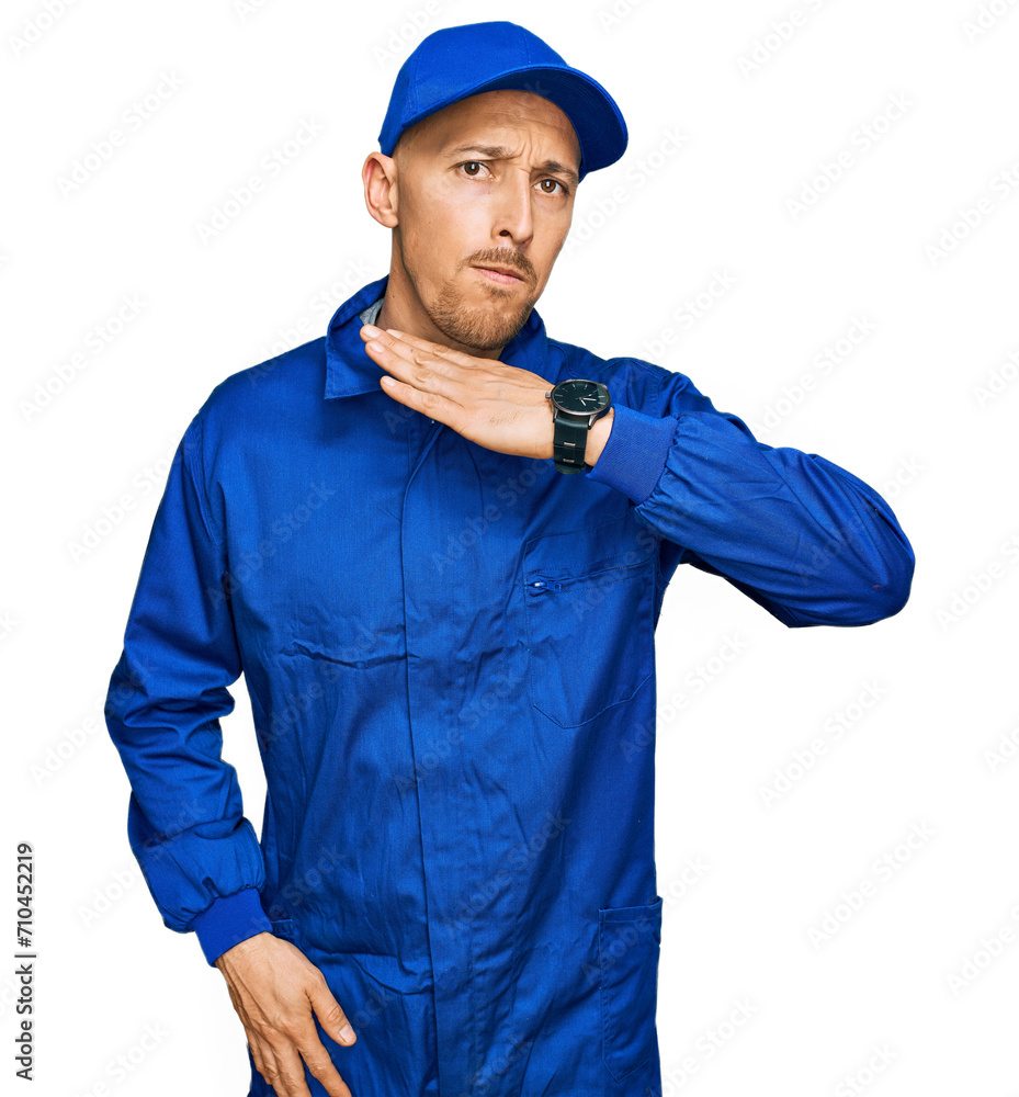 Bald man with beard wearing builder jumpsuit uniform cutting throat with hand as knife, threaten aggression with furious violence