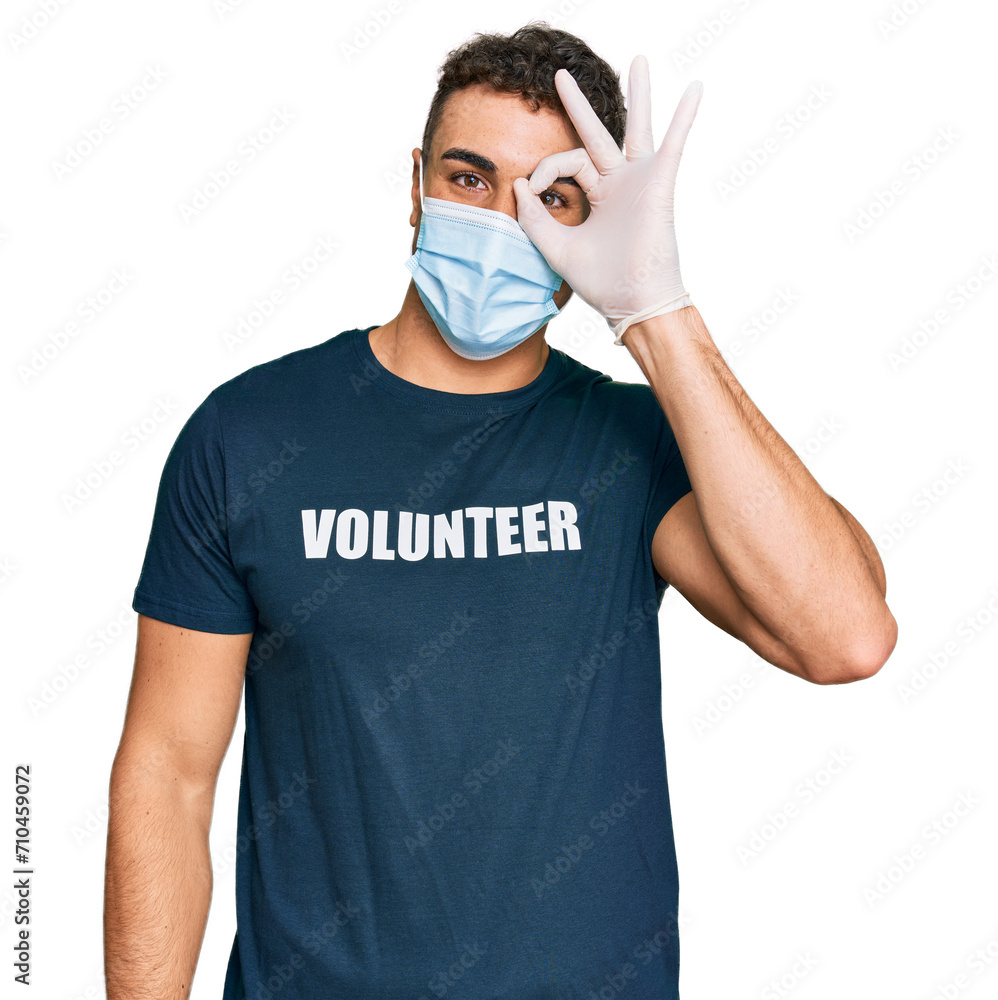 Hispanic young man wearing medical mask and volunteer t shirt smiling happy doing ok sign with hand on eye looking through fingers Hispanic young man wearing medical mask and volunteer t shirt smiling happy doing ok sign with hand on eye looking through fingers