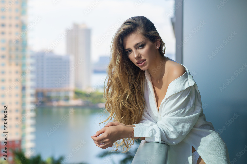 Woman on the balcony enjoying city view. Attractive girl with long blonde hair in shirt on balcony in the morning in city. Woman lifestyle in summer holiday vacation resting in hotel balcony.