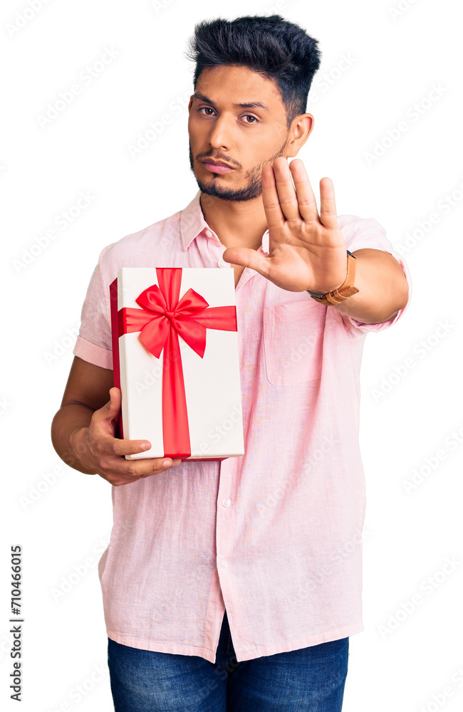 Handsome latin american young man holding gift with open hand doing stop sign with serious and confident expression, defense gesture Handsome latin american young man holding gift with open hand doing stop sign with serious and confident expression, defense gesture