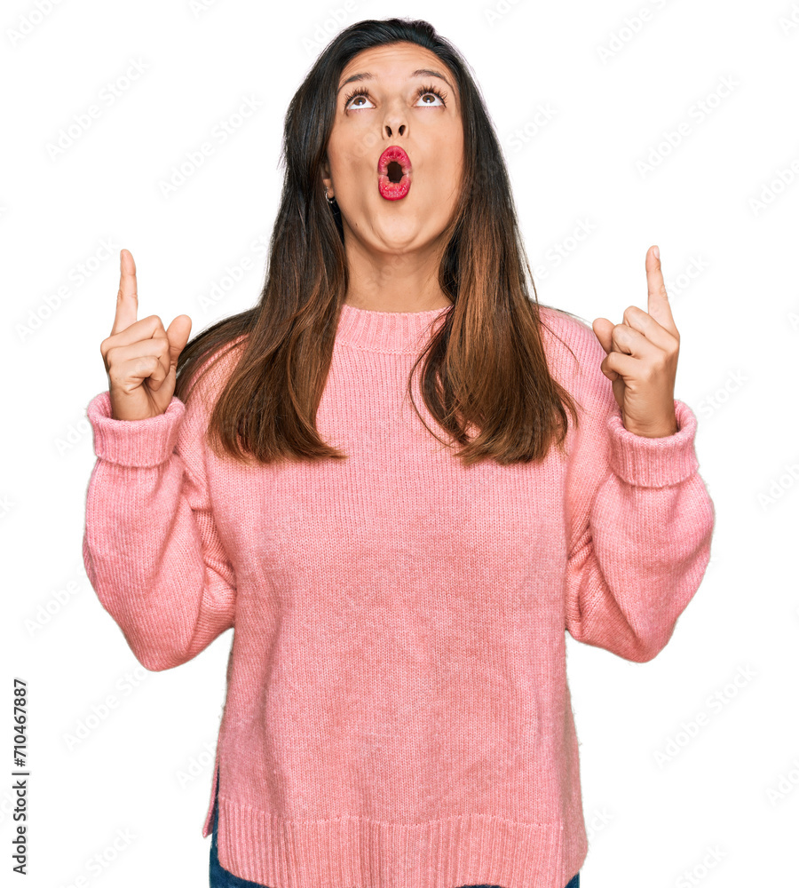 Beautiful hispanic woman wearing casual winter sweater amazed and surprised looking up and pointing with fingers and raised arms. Beautiful hispanic woman wearing casual winter sweater amazed and surprised looking up and pointing with fingers and raised arms.