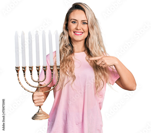 Young blonde woman holding menorah hanukkah jewish candle smiling happy pointing with hand and finger photo