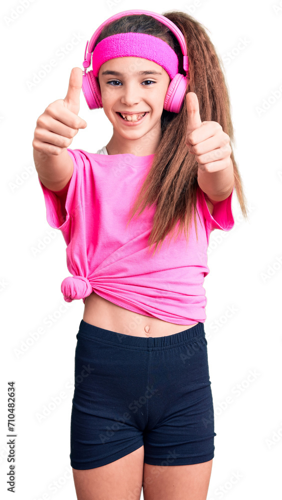 Cute hispanic child girl wearing gym clothes and using headphones approving doing positive gesture with hand, thumbs up smiling and happy for success. winner gesture. Cute hispanic child girl wearing gym clothes and using headphones approving doing positive gesture with hand, thumbs up smiling and happy for success. winner gesture.