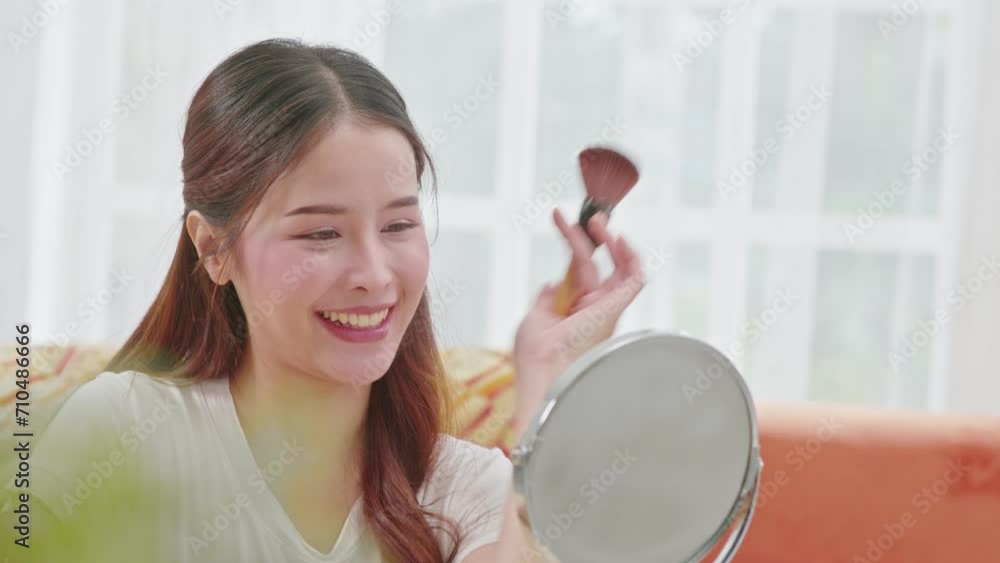 Happy Asian young woman smile and looking in mirror doing skin care applying make up brush on her cheek and beauty treatment at home,Wellness woman self care with beauty product at home